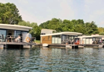 Modern houseboats are moored on a calm lake, surrounded by lush green trees and a clear blue sky.