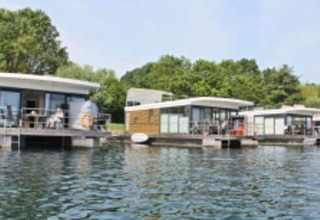 Moderne Hausboote liegen an einem ruhigen Gewässer, umgeben von üppigem grünen Wald und blauem Himmel.