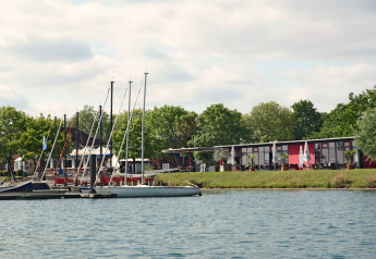 Foto van een houseboat aan de kade met meerdere zeilboten en moderne gebouwen tussen de bomen.