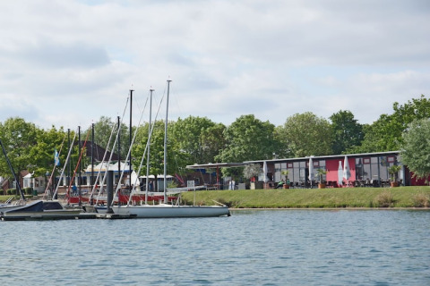 Foto von Hausboot am Ufer mit mehreren Segelbooten, modernen Gebäuden und grüner Baumlandschaft.