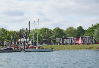 Photo of a houseboat dock with several sailboats and modern waterfront buildings surrounded by trees.