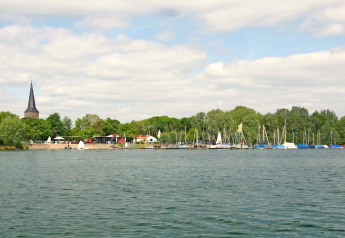 Uitzicht vanaf een woonboot op een meer met zeilboten, bomen en een kerktoren in de verte.