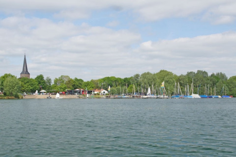 Blick von einem Hausboot auf einen See mit Segelbooten, Bäumen und einem Kirchturm im Hintergrund.