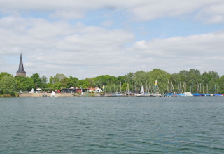 Blick von einem Hausboot auf einen See mit Segelbooten, Bäumen und einem Kirchturm im Hintergrund.