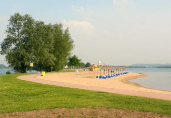 Rustig uitzicht op een zandstrand met blauwe stoelen, groen gras en kalm water onder een heldere lucht.