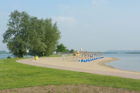 Ruhige Strandlandschaft mit blauen Strandkörben, Sand, ruhigem Wasser und grüner Wiese im Vordergrund.
