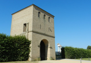 Foto eines alten Steintors mit Turm an einem klaren blauen Himmel, umgeben von Hecken und Sandweg.