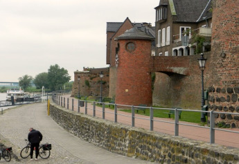 Rivierpromenade met fietsers, een aangemeerde boot en historische bakstenen gebouwen op de achtergrond.
