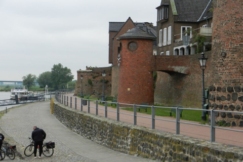 River promenade with cyclists, a boat docked at the pier, and historic brick buildings in the background.