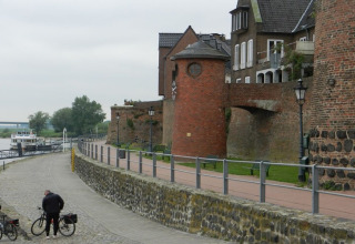 Flusspromenade mit Radfahrern, ein Boot am Ufer und historische Backsteinmauern im Hintergrund.