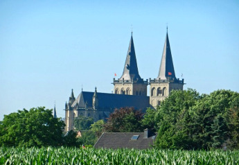 Foto van een grote kerk met twee torens achter bomen en een huisdak onder een heldere hemel.