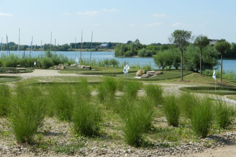 Blick auf einen Minigolfplatz am Seeufer mit grüner Bepflanzung, Bäumen und Wasser im Hintergrund.