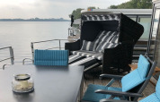 Outdoor seating area on a houseboat in Germany, with blue chairs, table, and striped lounger by the lake.