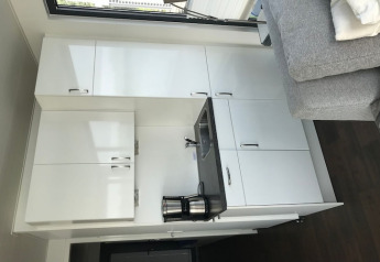 Modern compact kitchen on a houseboat with white cabinets, gray sofa, sink, and coffee maker by window.