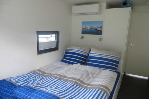Lichte slaapkamer op een woonboot met blauw-wit beddengoed, klein raam en een schilderij aan de muur.