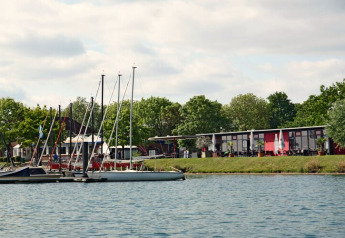 Woonboten en zeilbootjes aan een kalme rivier, omgeven door bomen en een modern gebouw op de oever.