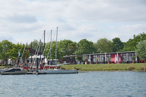 Houseboat e piccole barche a vela ormeggiate su un fiume tranquillo con alberi verdi e edifici moderni.