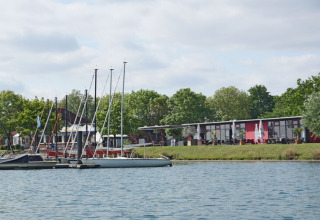 Woonboten en zeilbootjes aan een kalme rivier, omgeven door bomen en een modern gebouw op de oever.
