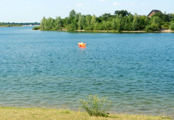 Foto van een meer met een oranje papieren bootje, groene bomen achteraan en een huis zichtbaar.