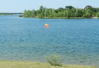Foto von einem ruhigen See mit einem orangefarbenen Papierboot und einem Haus im Hintergrund zwischen Bäumen.