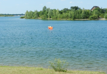 Photo d'un lac avec un bateau en papier orange flottant, arbres verts en arrière-plan et une maison.