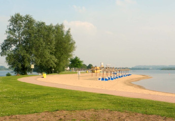 Zandstrand aan het water met blauwe ligzetels en rieten parasols, omringd door gras en bomen.