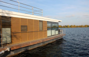 Moderne woonboot met houten lamellen en grote ramen drijvend op een rustig meer onder een heldere lucht.