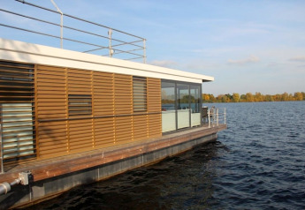 Moderne woonboot met houten lamellen en grote ramen drijvend op een rustig meer onder een heldere lucht.