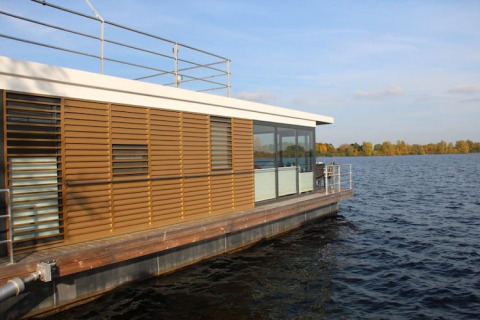 Casa galleggiante moderna con persiane in legno e ampie vetrate, ormeggiata su un lago sotto cielo sereno.