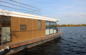 Maison flottante moderne en bois et grandes vitres, flottant sur un lac calme sous ciel bleu clair et paisible.