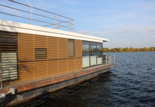 Moderne woonboot met houten lamellen en grote ramen drijvend op een rustig meer onder een heldere lucht.