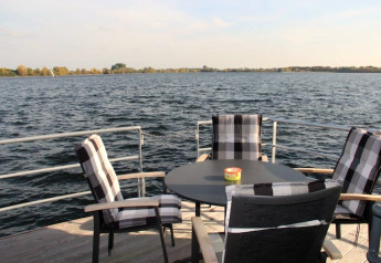 Uitzicht vanaf een woonboot met vier zwart-wit stoelen rond een tafel, uitkijkend over het water.