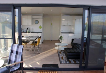 Uitzicht door grote glazen deuren op een modern woonbootinterieur met keuken en zithoek.