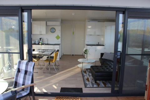 Vista attraverso grandi porte in vetro su un moderno interno di houseboat con cucina e zona giorno.