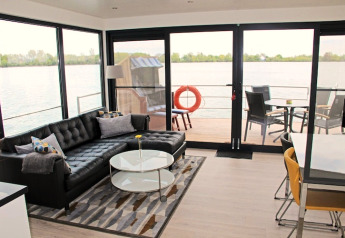 Woonkamer op een woonboot met zwarte bank, salontafel en uitzicht op het water door grote ramen.