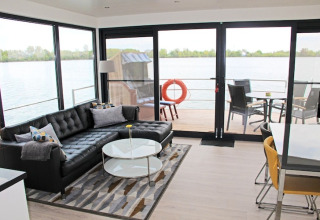 Woonkamer op een woonboot met zwarte bank, salontafel en uitzicht op het water door grote ramen.