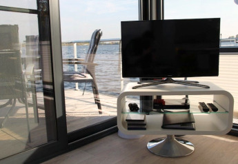 Woonkamer op een woonboot met moderne tv, uitzicht op het water en terras met stoelen buiten.