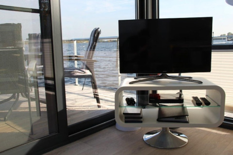 Woonkamer op een woonboot met moderne tv, uitzicht op het water en terras met stoelen buiten.