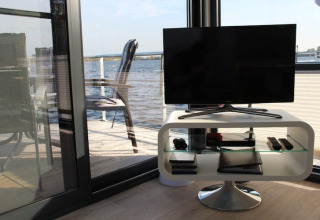 Soggiorno di una houseboat con TV moderna e vista sull’acqua attraverso ampie porte in vetro.