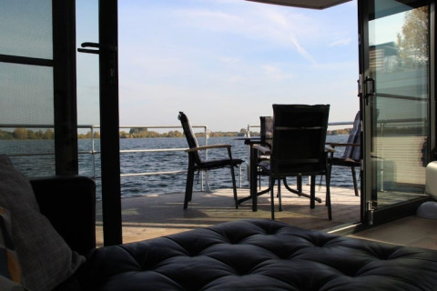 Uitzicht vanuit een woonboot door openstaande deuren op een terras met tafel en stoelen aan het water.