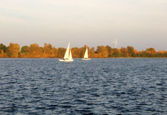 Uitzicht vanaf een woonboot over een meer met twee zeilboten en herfstbomen langs de oever.