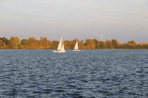 View from a houseboat overlooking a lake with two sailboats and autumn trees lining the shore.