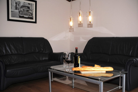 Woonkamer in chalet Waldferienpark Gerolstein 2 in Duitsland met zwarte zetels, wijn en baguettebrood.