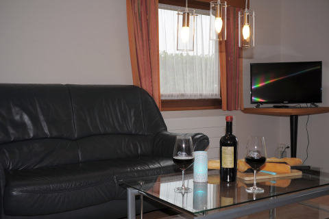Gezellige woonkamer in Waldferienpark Gerolstein 2 met zwarte sofa, wijn, tv en vers stokbrood.
