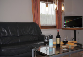 Gezellige woonkamer in Waldferienpark Gerolstein 2 met zwarte sofa, wijn, tv en vers stokbrood.