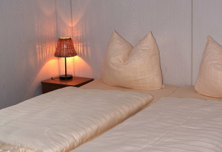 Slaapkamer met twee bedden, zachte kussens, lichte dekens en een brandende lamp in chalet Waldferienpark.
