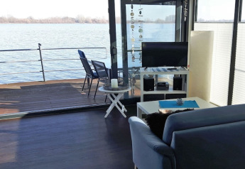 Living area on houseboat with sofa, TV and deck overlooking water, seen at Floating 44 in Germany.