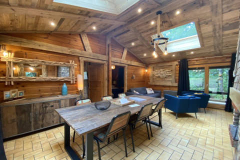 Interno di chalet rustico in legno con tavolo da pranzo, divani blu, lucernario e vista sul bosco in Francia.