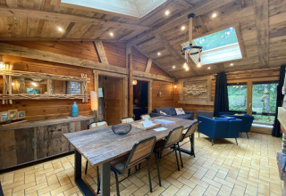 Rustiek houten chaletinterieur met eettafel, blauwe banken, dakraam en boszicht in Frankrijk.