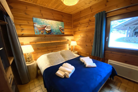 Slaapkamer in het houten chalet LE FISHING LODGE in Frankrijk, met houten muren, raam en blauw dekbed.